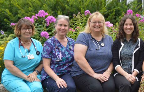 Four women, some in scrubs with stethoscopes, sit smiling in front of blooming pink flowers outdoors.
