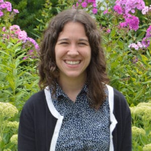 Smiling woman with curly hair stands in front of blooming pink and green flowers outdoors.