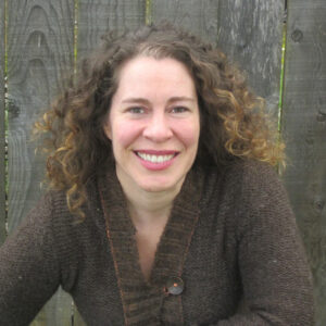 Smiling woman with curly hair wearing a brown sweater, sitting in front of a wooden fence. Audrey Wehmeyer, Smiling woman with curly hair wearing a brown sweater, sitting in front of a wooden fence.