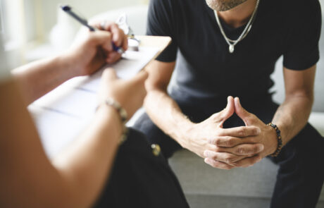 A person writes on a clipboard during a consultation with a man sitting opposite them.